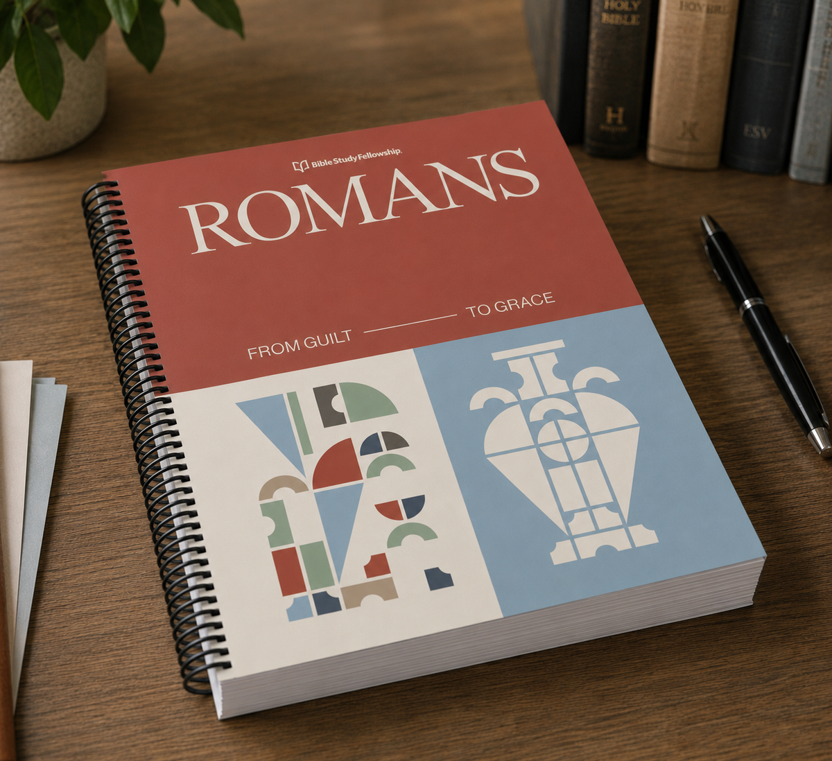 A spiral-bound book titled 'ROMANS' from the Bible Technology series, featuring a terracotta red top half and a light blue bottom half with white typography and abstract geometric illustrations. The book is photographed on a wooden desk next to a pen, with books and a plant blurred in the background.
