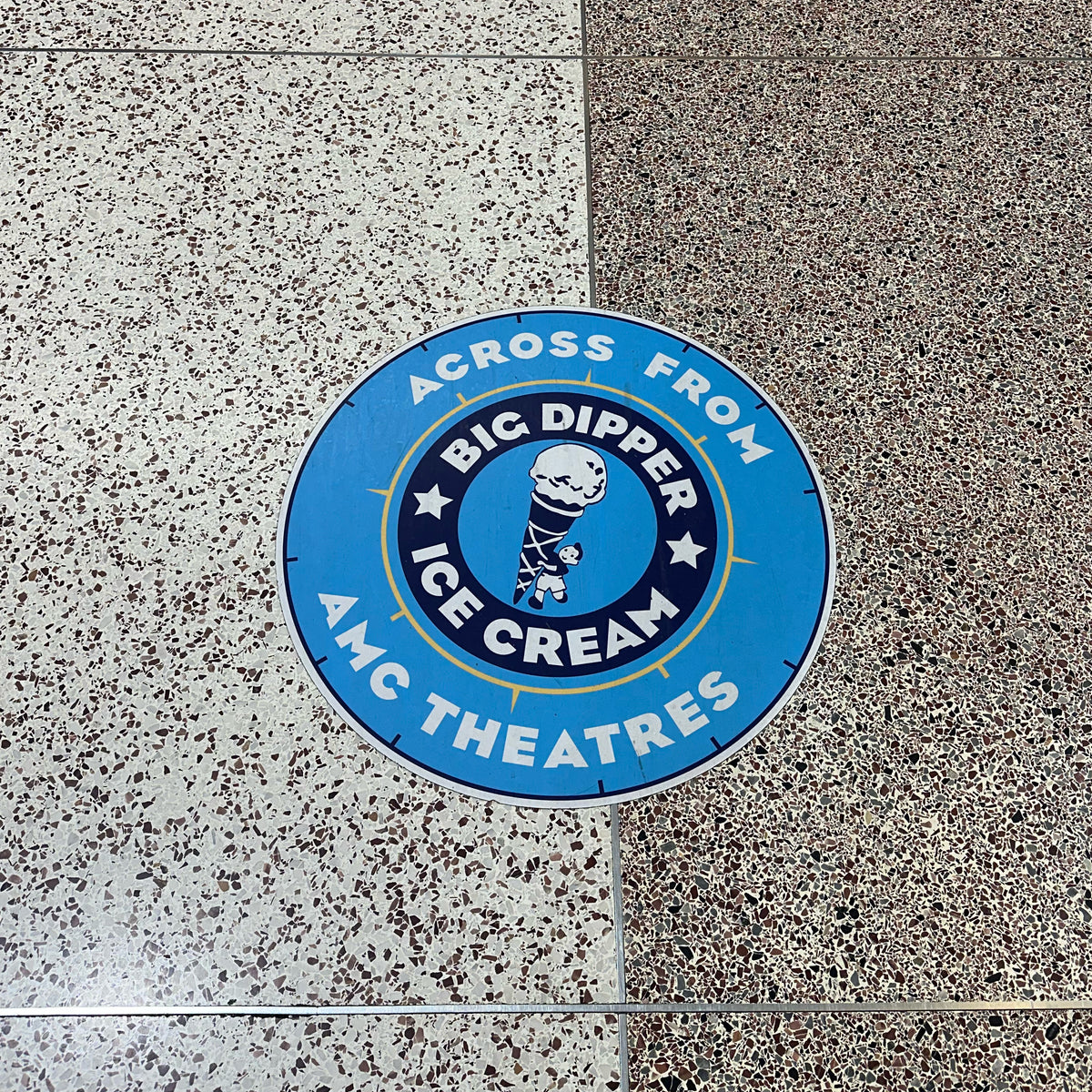 A circular blue custom floor graphic placed on a speckled floor surface, with white text and graphics describing an ice cream brand relative to AMC Theatres.
