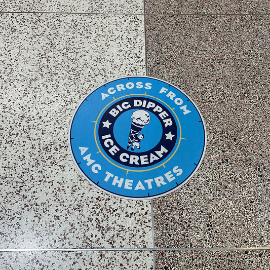 A circular blue custom floor graphic placed on a speckled floor surface, with white text and graphics describing an ice cream brand relative to AMC Theatres.