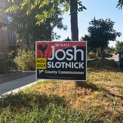 A political campaign yard sign on a lawn reading Re-elect Josh Slotnick County Commissioner Nov 2024, featuring a rooster weather vane logo on a black and red background next to a tree.
