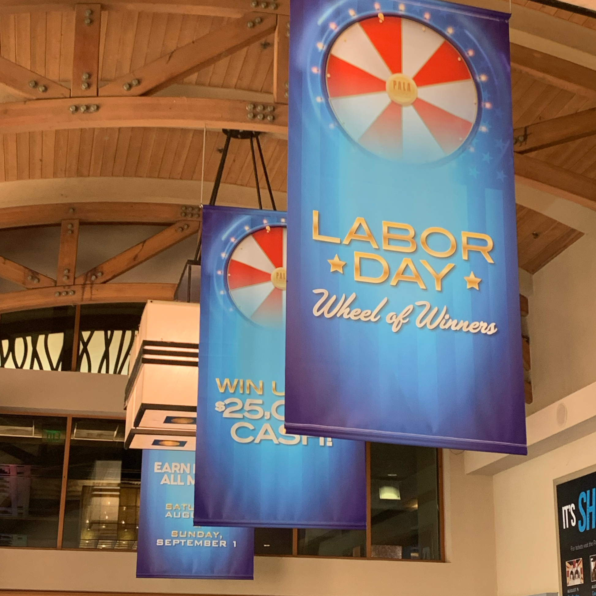 Three large indoor, promotional banners hung from the ceiling in a line reading "Labor Day Wheel of Fortune", showcasing the customizability of signage that is printable by 760 Print. 