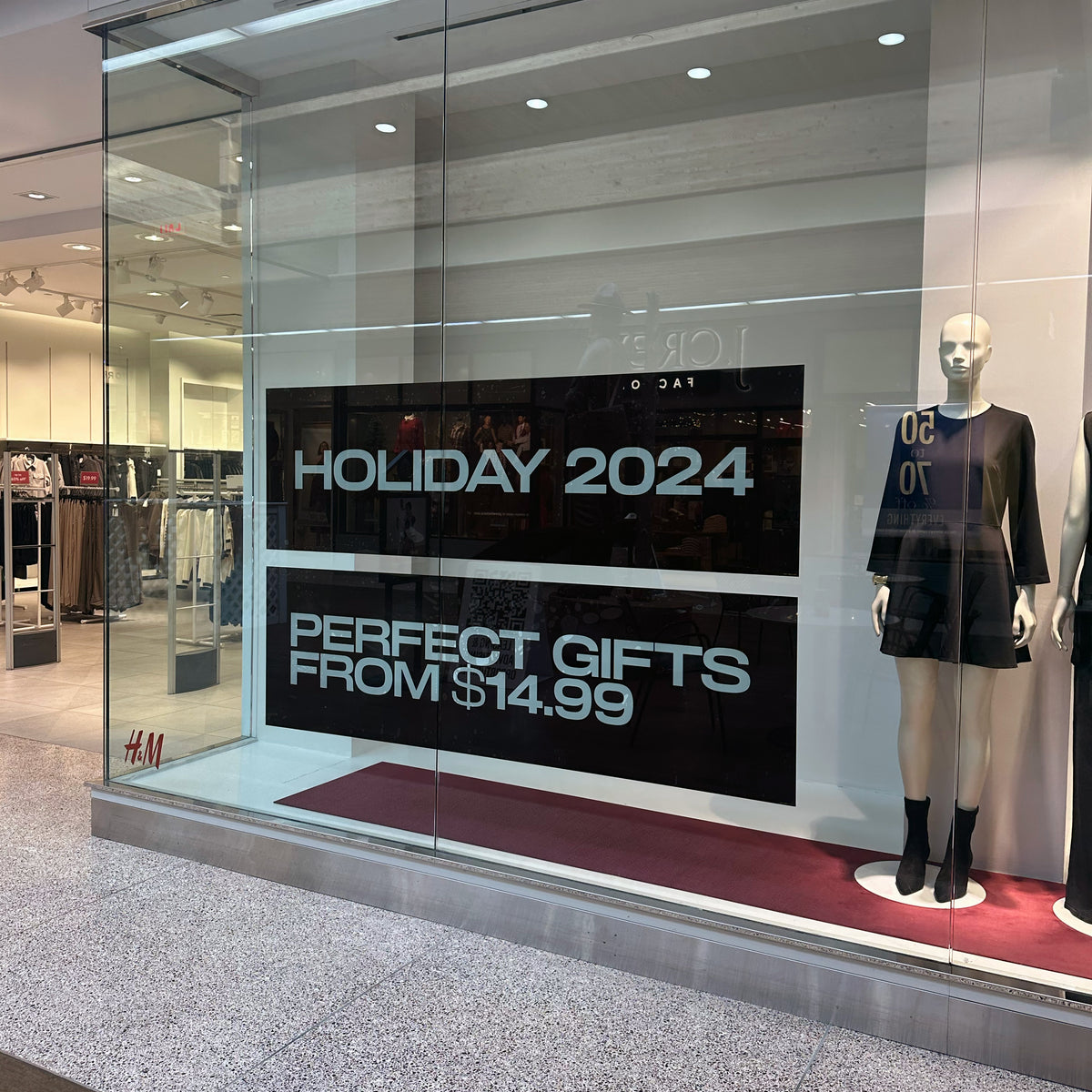 An H and M retail clothing store window display featuring two large black banners reading HOLIDAY 2024 and PERFECT GIFTS FROM 14.99 dollars next to a mannequin dressed in a black long-sleeve mini dress and ankle boots.