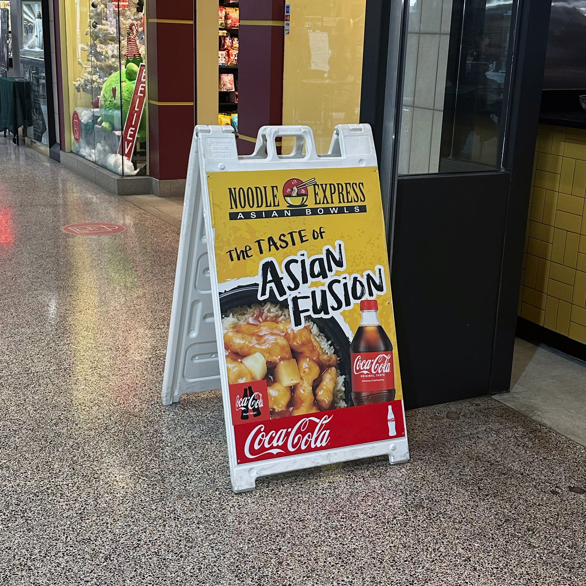 A custom printed white Plasticade A-frame sidewalk sign for Noodle Express Asian Bowls, manufactured by 760 Print and displayed in a shopping mall corridor.