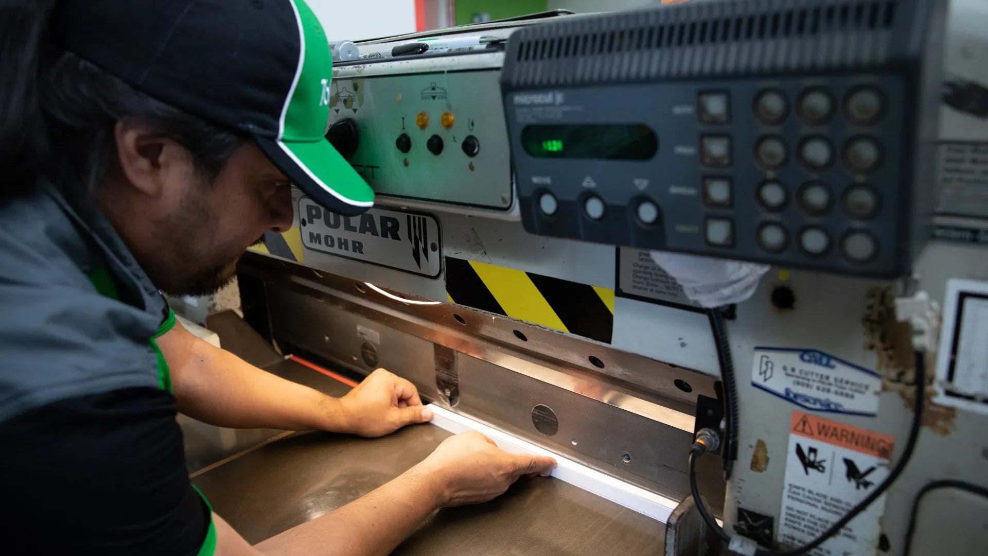 A man wearing a green and black baseball cap carefully aligns a stack of paper on the bed of a large industrial Polar Mohr paper cutter.