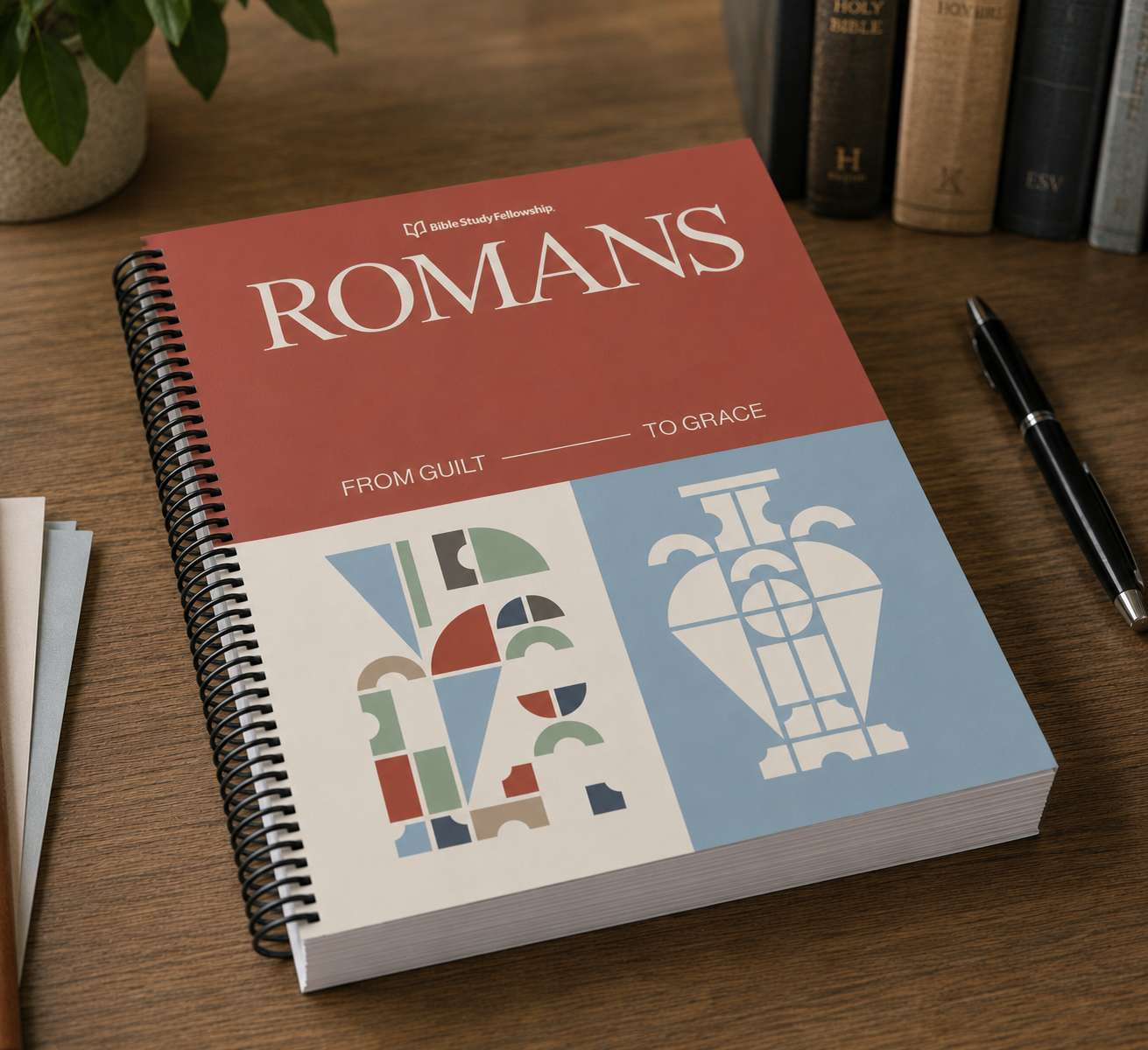 A spiral-bound book titled 'ROMANS' from the Bible Technology series, featuring a terracotta red top half and a light blue bottom half with white typography and abstract geometric illustrations. The book is photographed on a wooden desk next to a pen, with books and a plant blurred in the background.